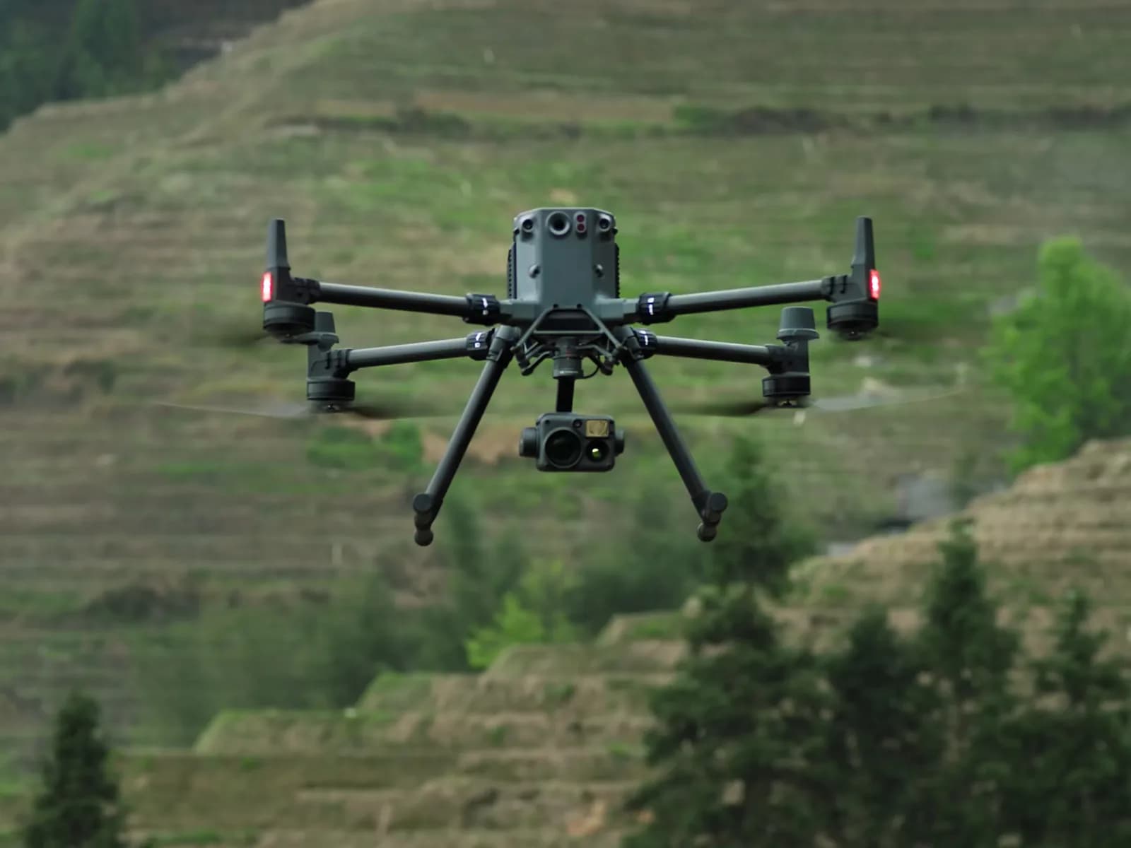 DJI Matrice 350 RTK en vol avec une nacelle caméra, devant une colline en terrasses
