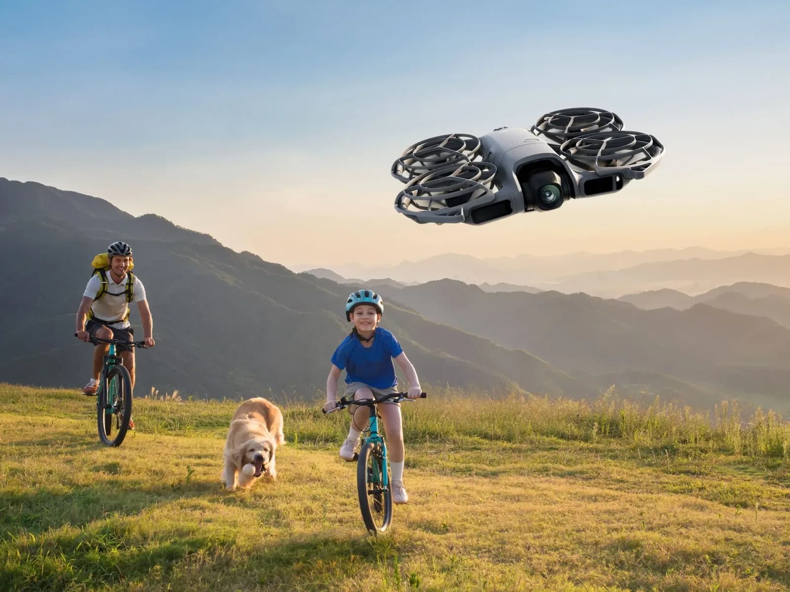 DJI Neo 2 suivant un enfant et un adulte à vélo accompagnés d'un chien dans une prairie de montagne