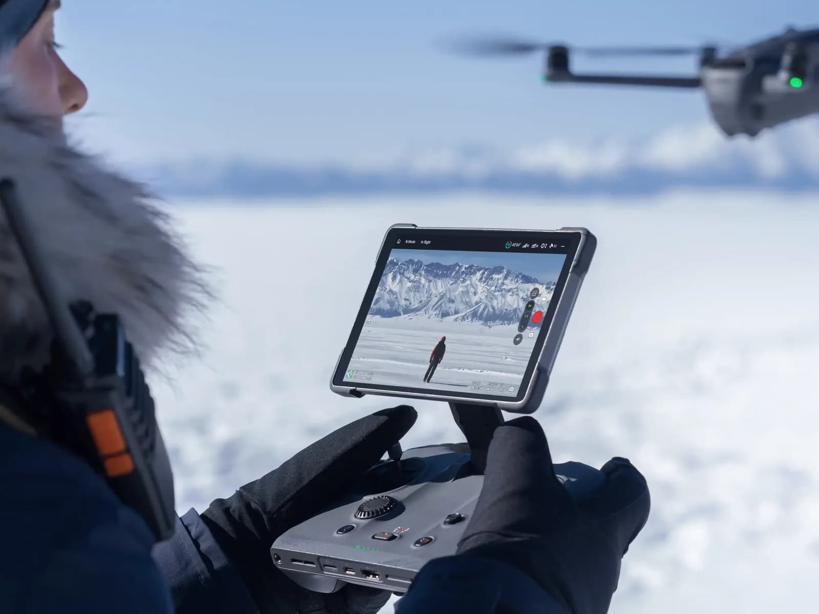 Télépilote en tenue grand froid utilisant la radiocommande DJI RC Pro 2 sur un terrain enneigé, drone en vol